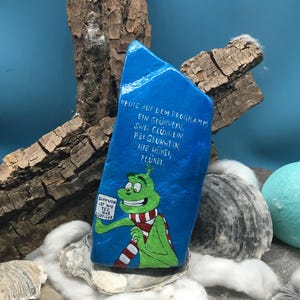 Pode incluir: Uma pedra azul pintada &agrave; m&atilde;o com texto em alem&atilde;o e uma personagem de desenho animado segurando uma placa. A pedra apresenta o texto "HEUTE AUF DEM PROGRAMM EIN GL&Uuml;HWEIN, ZWEI GL&Uuml;HWEIN. BEI GL&Uuml;HWEIN HIE H&Uuml;NEL, FL&Uuml;NEI."