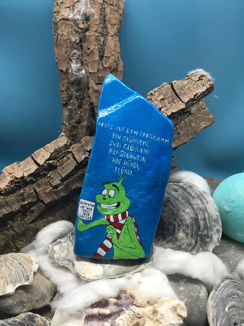 Pode incluir: Uma pedra azul pintada &agrave; m&atilde;o com texto em alem&atilde;o e uma personagem de desenho animado segurando uma placa. A pedra apresenta o texto "HEUTE AUF DEM PROGRAMM EIN GL&Uuml;HWEIN, ZWEI GL&Uuml;HWEIN. BEI GL&Uuml;HWEIN HIE H&Uuml;NEL, FL&Uuml;NEI."