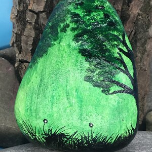 K&ouml;nnte beinhalten: Handbemalter Stein mit der Darstellung einer gr&uuml;nen Landschaft mit einer schwarzen Silhouette eines Baumes und wei&szlig;en Blumen.