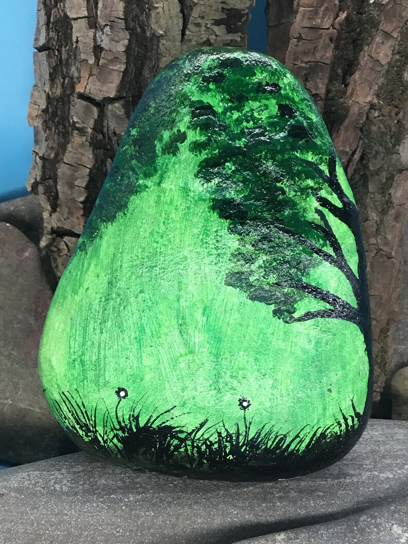 K&ouml;nnte beinhalten: Handbemalter Stein mit der Darstellung einer gr&uuml;nen Landschaft mit einer schwarzen Silhouette eines Baumes und wei&szlig;en Blumen.