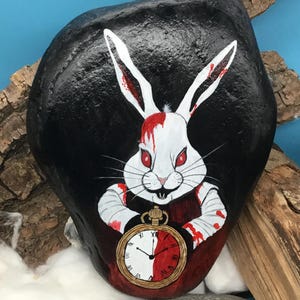 May include: Hand-painted rock art depicting a white rabbit with red eyes and blood-splattered fur and clothing. The rabbit holds a pocket watch with a red and white face. The background is black, with a blue backdrop and wood accents.