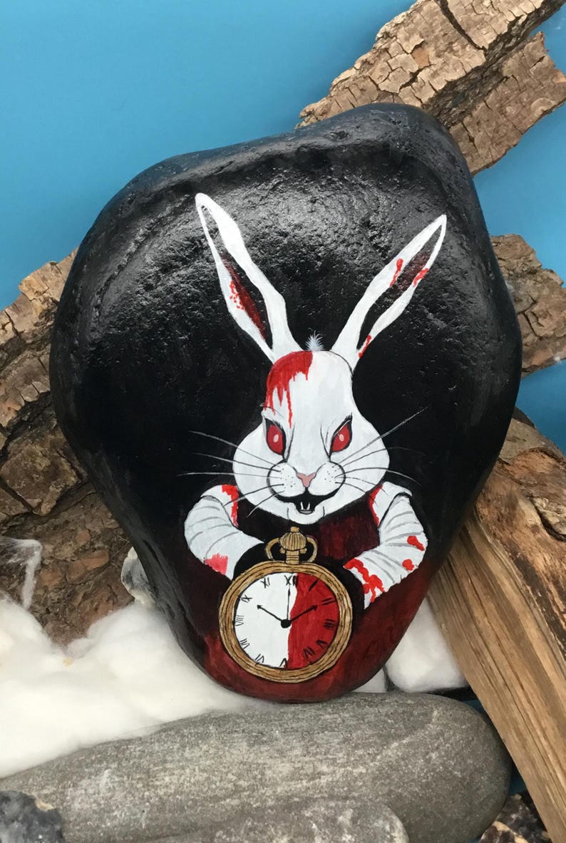 May include: Hand-painted rock art depicting a white rabbit with red eyes and blood-splattered fur and clothing. The rabbit holds a pocket watch with a red and white face. The background is black, with a blue backdrop and wood accents.