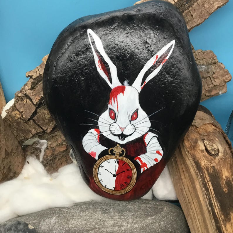 May include: A hand-painted rock features a white rabbit with red eyes and blood-splattered fur, holding a pocket watch. The rabbit is set against a black background. The rock is approximately 20 cm long.