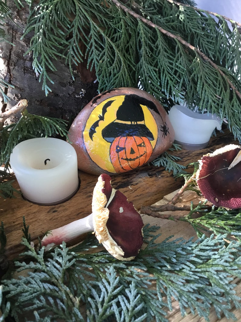 May include: A painted rock featuring a witch's hat and a jack-o'-lantern face. The rock is surrounded by green foliage and two white candles.