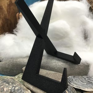 May include: A black metal display stand with a textured finish. The stand has an angled, open frame design, placed on grey stones and shells. The background features white fluffy material and wood.