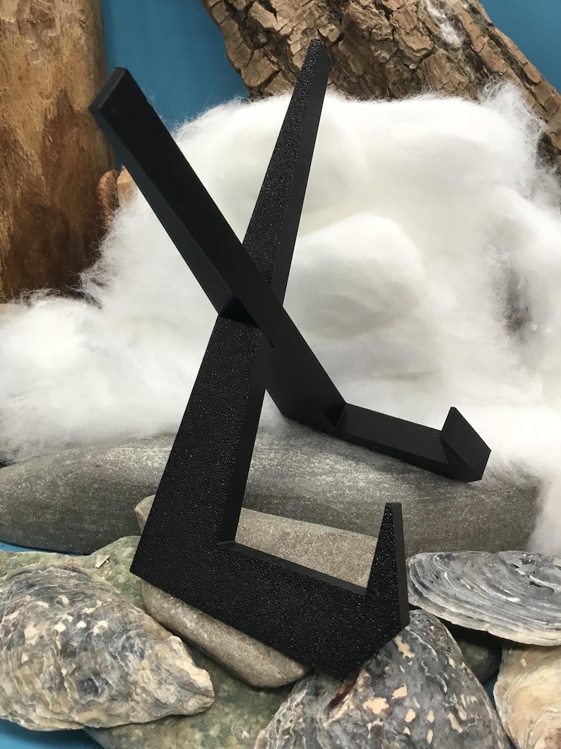 May include: A black metal display stand with a textured finish. The stand has an angled, open frame design, placed on grey stones and shells. The background features white fluffy material and wood.