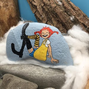 May include: A light blue painted rock featuring a hand-painted illustration of a character with bright red hair in pigtails, wearing a yellow dress, blue polka-dot shorts, and black boots with yellow and black striped socks, depicted in a seated pose.