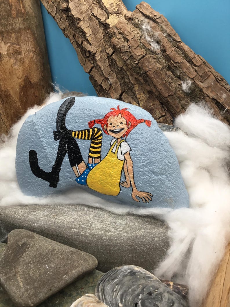 May include: A light blue painted rock featuring a hand-painted illustration of a character with bright red hair in pigtails, wearing a yellow dress, blue polka-dot shorts, and black boots with yellow and black striped socks, depicted in a seated pose.
