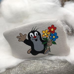 May include: A hand-painted, gray stone features a cartoon mole holding a bouquet of flowers. The mole is black, gray, and tan with a red nose and a cheerful expression. The flowers are red, yellow, and blue.