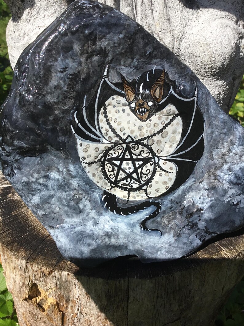 May include: Hand-painted rock art featuring a black and white bat with a pentagram design. The bat has brown ears and a menacing expression. The rock is painted in shades of gray and black, with a textured appearance. A unique piece of gothic home decor.