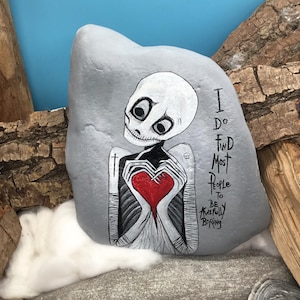 May include: A hand-painted gray rock features a skeleton holding a red heart. The text "I do find most people to be awfully boring" is written on the rock. The artwork is set against a blue background.