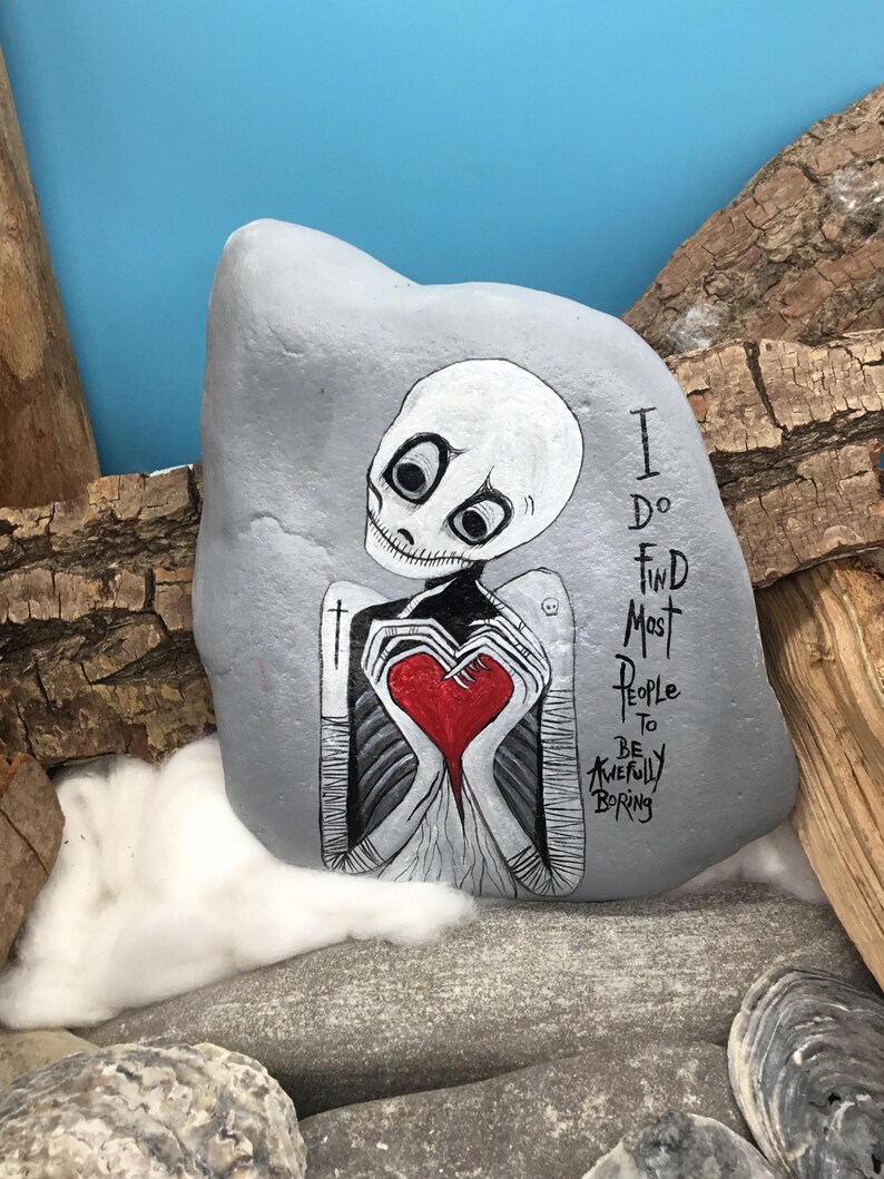 May include: A hand-painted gray rock features a skeleton holding a red heart. The text "I do find most people to be awfully boring" is written on the rock. The artwork is set against a blue background.