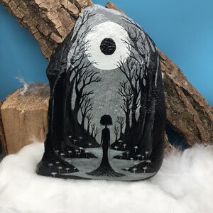 May include: Hand-painted rock art depicting a grayscale forest scene with a full moon and a silhouetted figure. The artwork features detailed trees and a serene, dreamlike quality. The rock is displayed on a bed of white fluff.