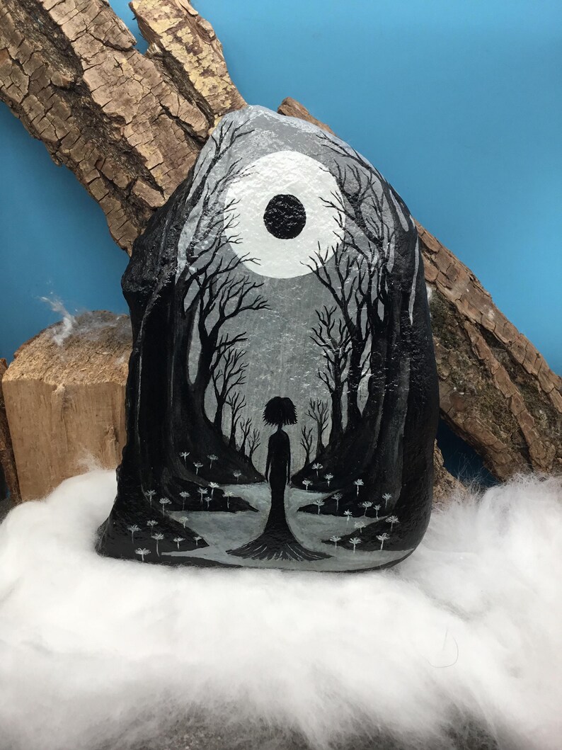 May include: Hand-painted rock art depicting a grayscale forest scene with a full moon and a silhouetted figure. The artwork features detailed trees and a serene, dreamlike quality. The rock is displayed on a bed of white fluff.