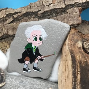 May include: Hand-painted rock art featuring a cartoon character in a black robe and green tie, holding a wand. The character has white hair and green eyes. The rock is gray and set against a backdrop of wood and blue.