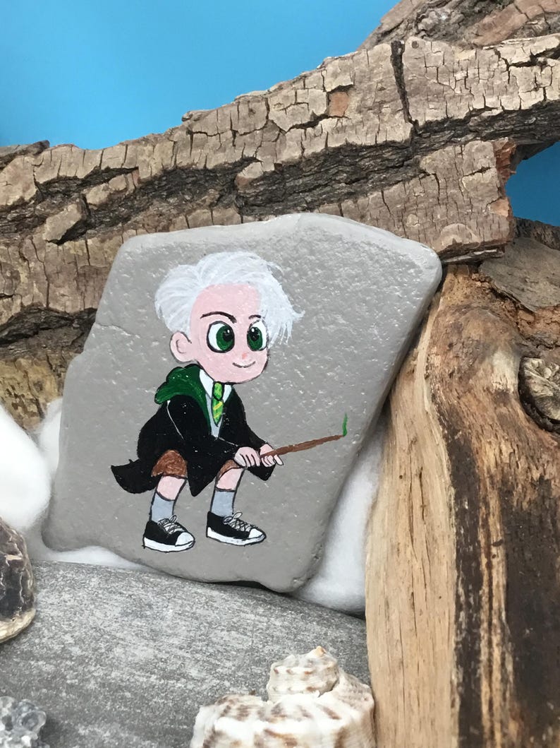 May include: Hand-painted rock art featuring a cartoon character in a black robe and green tie, holding a wand. The character has white hair and green eyes. The rock is gray and set against a backdrop of wood and blue.