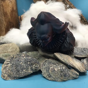 May include: A dark, stylized sculpture of a figure with a ruffled collar and large ears, possibly a character from a fantasy setting. The sculpture is placed among rocks and oyster shells, with a backdrop of cotton and wood against a blue surface.