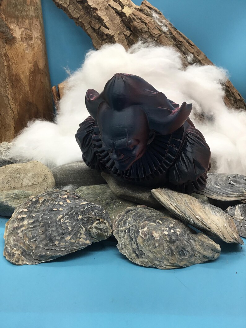 May include: A dark, stylized sculpture of a figure with a ruffled collar and large ears, possibly a character from a fantasy setting. The sculpture is placed among rocks and oyster shells, with a backdrop of cotton and wood against a blue surface.