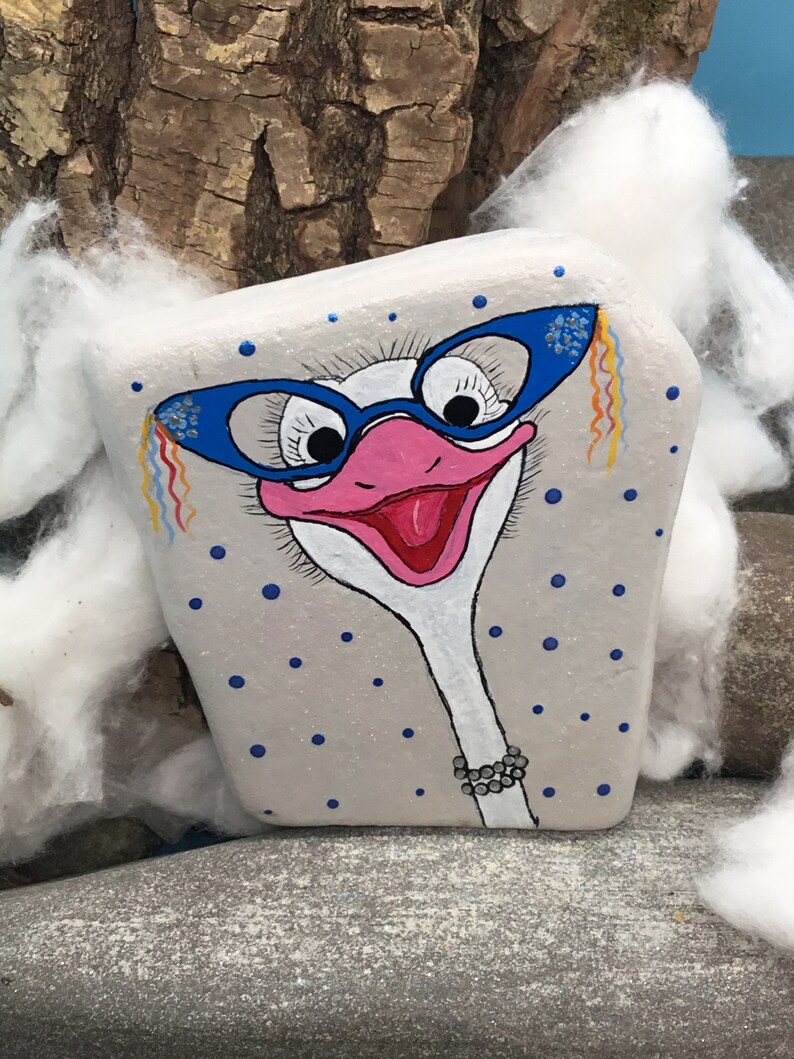May include: A painted rock with a white background and blue polka dots. The rock depicts an ostrich with blue glasses, a pink beak, and a black beaded necklace.