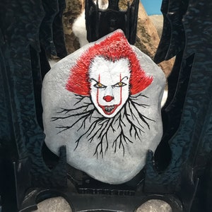 May include: Hand-painted rock art featuring a scary clown with red hair, white face paint, and black roots. The clown has a menacing expression with sharp teeth and yellow eyes.