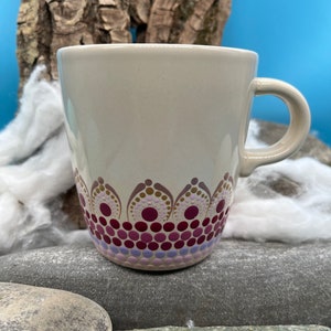 Puede incluir: Una taza de cerámica blanca con un diseño de mandala pintado a mano en tonos de morado, rosa y dorado. El diseño presenta una serie de círculos concéntricos y puntos.