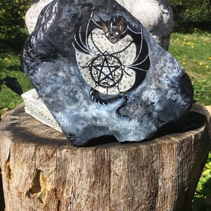 May include: A hand-painted, dark gray rock with a bat and pentagram design. The bat has black wings and a tan face, and is surrounded by a white dotted pattern. A black pentagram is in the center.