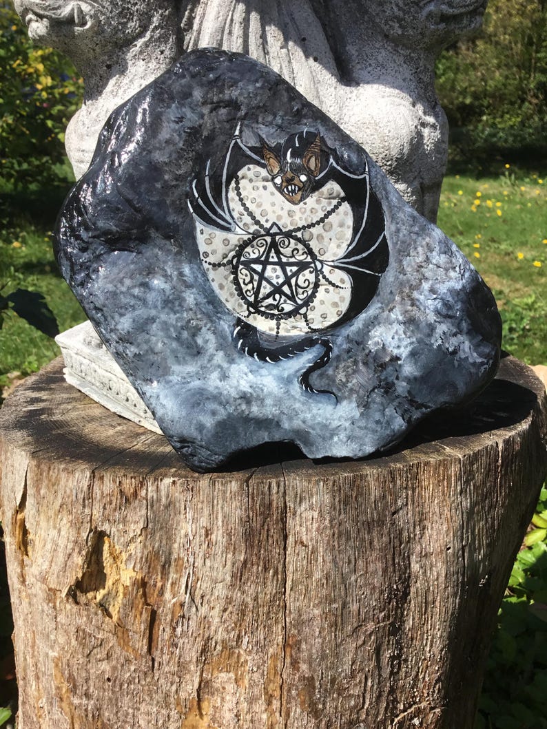 May include: A hand-painted, dark gray rock with a bat and pentagram design. The bat has black wings and a tan face, and is surrounded by a white dotted pattern. A black pentagram is in the center.