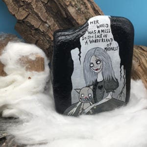 May include: A hand-painted black rock featuring a gothic illustration of a pale woman with gray hair and a doll. The text reads "Her world was a mess so she lost in a wonderland of madness."