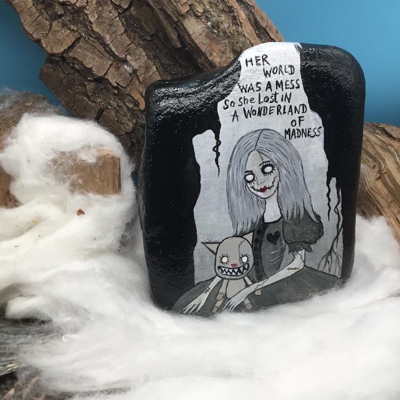 May include: A hand-painted black rock featuring a gothic illustration of a pale woman with gray hair and a doll. The text reads "Her world was a mess so she lost in a wonderland of madness."