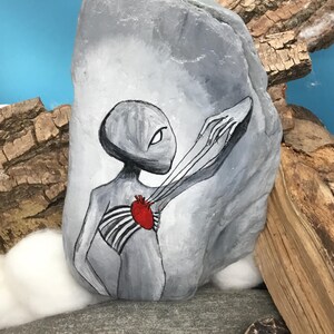 K&ouml;nnte beinhalten: Ein handbemalter grauer Stein zeigt eine Alienfigur mit einem roten Herzen und Brustkorb. Der Au&szlig;erirdische streckt einen Arm aus. Der Hintergrund ist hellblau mit Holz und anderen Steinen.