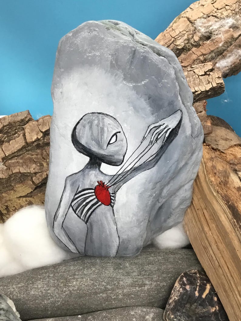 K&ouml;nnte beinhalten: Ein handbemalter grauer Stein zeigt eine Alienfigur mit einem roten Herzen und Brustkorb. Der Au&szlig;erirdische streckt einen Arm aus. Der Hintergrund ist hellblau mit Holz und anderen Steinen.