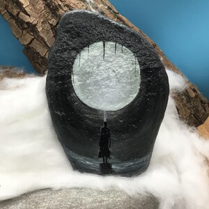 Pu&ograve; includere: Una roccia nera dipinta con un cerchio bianco e una silhouette. La figura &egrave; in piedi nell'acqua sotto il cerchio, con linee verticali che scendono dal cerchio. La roccia &egrave; esposta con legno e lanugine bianca.