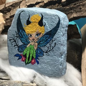 May include: Hand-painted rock art featuring a fairy with blue wings, green clothing, and yellow hair. The fairy is sitting on a pink flower. The rock is painted a light blue color.