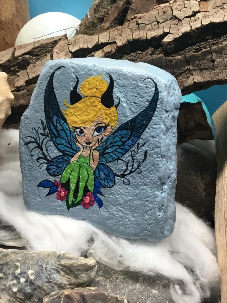 May include: Hand-painted rock art featuring a fairy with blue wings, green clothing, and yellow hair. The fairy is sitting on a pink flower. The rock is painted a light blue color.