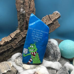 Pode incluir: Uma pedra azul pintada &agrave; m&atilde;o com texto em alem&atilde;o e um personagem de desenho animado com um cachecol vermelho e branco. A pedra, com as palavras "Gl&uuml;hwein" e "Fl&uuml;nei", repousa entre conchas e pedras. O personagem segura uma placa.