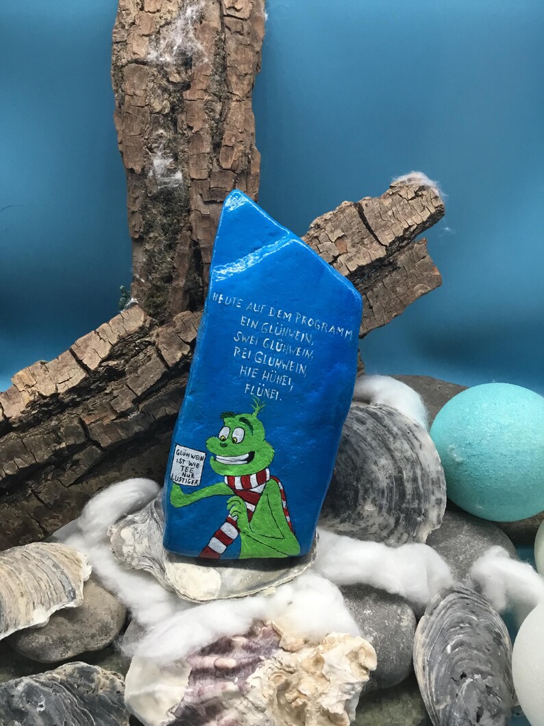 Pode incluir: Uma pedra azul pintada &agrave; m&atilde;o com texto em alem&atilde;o e um personagem de desenho animado com um cachecol vermelho e branco. A pedra, com as palavras "Gl&uuml;hwein" e "Fl&uuml;nei", repousa entre conchas e pedras. O personagem segura uma placa.