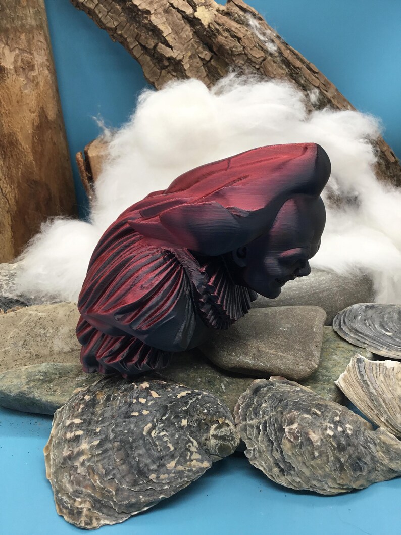 May include: A burgundy and black bust sculpture of a person wearing a ruffled collar and hat. The sculpture is resting on a bed of rocks and seashells, with a backdrop of wood and cotton. The bust is a decorative art piece.