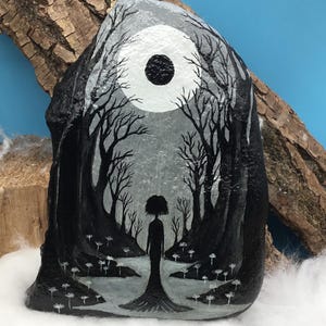 May include: Hand-painted rock art featuring a surreal forest scene. The artwork depicts a silhouetted figure standing in a path surrounded by trees, under a moon. The color palette is primarily black, white, and gray, creating a moody and artistic aesthetic.