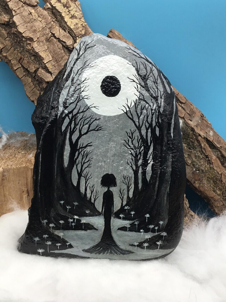 May include: Hand-painted rock art featuring a surreal forest scene. The artwork depicts a silhouetted figure standing in a path surrounded by trees, under a moon. The color palette is primarily black, white, and gray, creating a moody and artistic aesthetic.