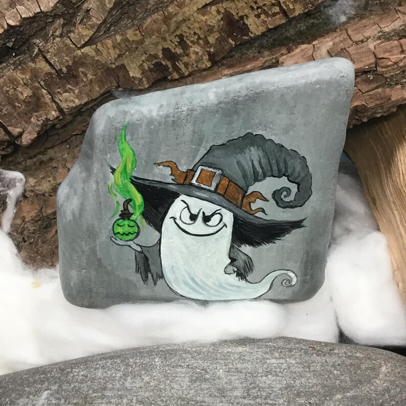 Peut inclure: Une pierre grise peinte &agrave; la main repr&eacute;sente un fant&ocirc;me de dessin anim&eacute; portant un chapeau de sorci&egrave;re avec une boucle marron et tenant une potion verte. Le fant&ocirc;me a un sourire espi&egrave;gle et est entour&eacute; d'une vapeur verte tourbillonnante. L'&oelig;uvre est sur fond naturel.