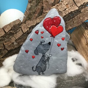 May include: A hand-painted gray rock with a cartoon dog holding two red heart-shaped balloons. The rock is surrounded by smaller red hearts.