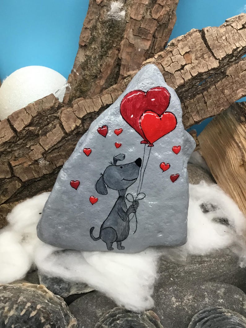 May include: A hand-painted gray rock with a cartoon dog holding two red heart-shaped balloons. The rock is surrounded by smaller red hearts.