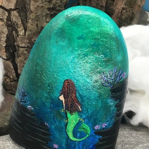 May include: Hand-painted rock art depicting a mermaid with long brown hair and a green tail, swimming in a turquoise blue ocean with black rocks and purple coral.