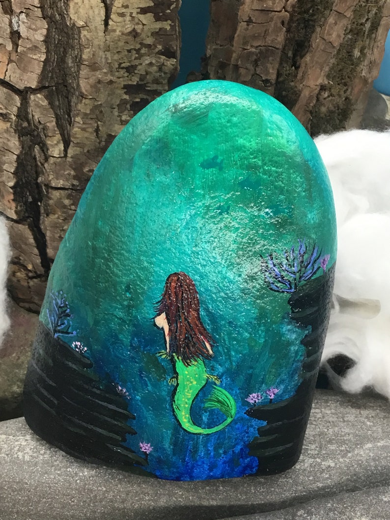 May include: Hand-painted rock art depicting a mermaid with long brown hair and a green tail, swimming in a turquoise blue ocean with black rocks and purple coral.