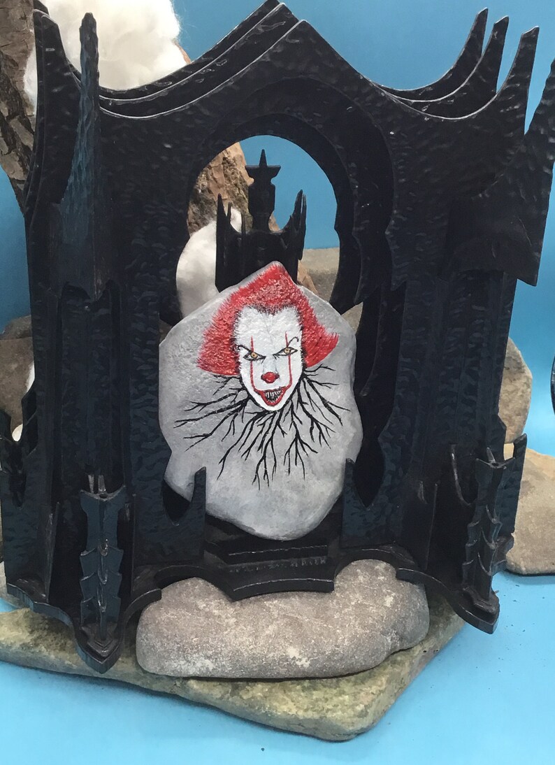 May include: A black metal frame with a painted stone inside. The stone is painted with a scary clown face with red hair and a white background. The clown has black eyes and a red nose.