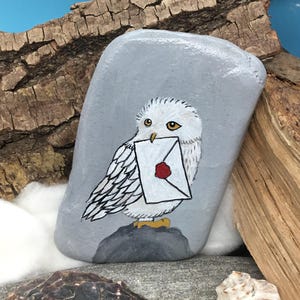 May include: A hand-painted, grey stone features a white owl holding a letter with a red wax seal. The owl is perched on a grey rock, with a backdrop of wood and a blue sky. This decorative stone is a unique art piece.