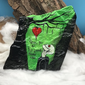 May include: Hand-painted rock art featuring a green and black design with a red heart and a stylized figure. The text "Thank You For This Wonderful Painful Life" is included. The artwork is set against a backdrop of wood and cotton.