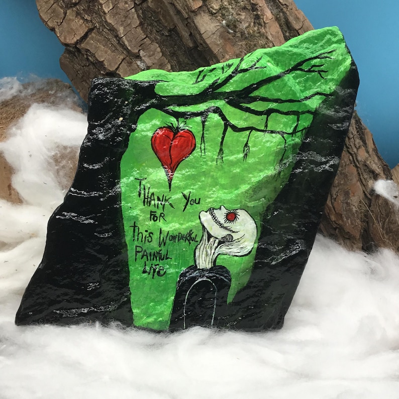 May include: Hand-painted rock art featuring a green and black design with a red heart and a stylized figure. The text "Thank You For This Wonderful Painful Life" is included. The artwork is set against a backdrop of wood and cotton.