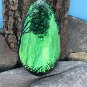 K&ouml;nnte beinhalten: Ein handbemalter Stein mit gr&uuml;nem Hintergrund und einer schwarzen Silhouette eines Baumes. Der Stein ist glatt und oval.