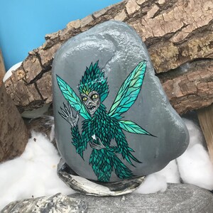 May include: A hand-painted, gray stone features a detailed illustration of a fairy. The fairy has green and teal wings, a matching feathered outfit, and a stern expression. The artwork is set against a backdrop of wood and cotton.